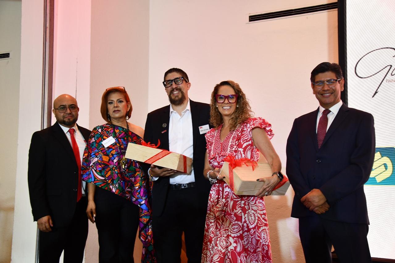 Cinco personas en el escenario; dos de ellas sostienen reconocimientos de madera con moño rojo