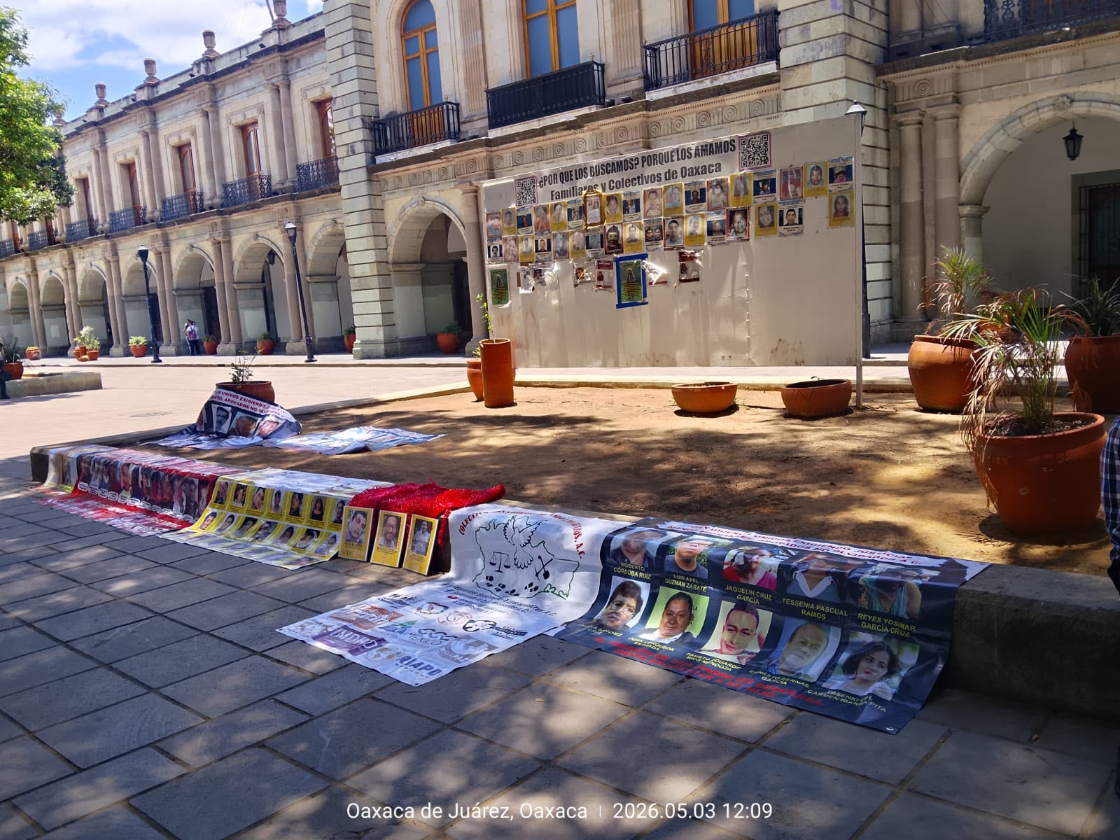 Lonas, carteles y fotografías de personas desaparecidas tendidas en el piso de una plaza, frente a un edificio colonial